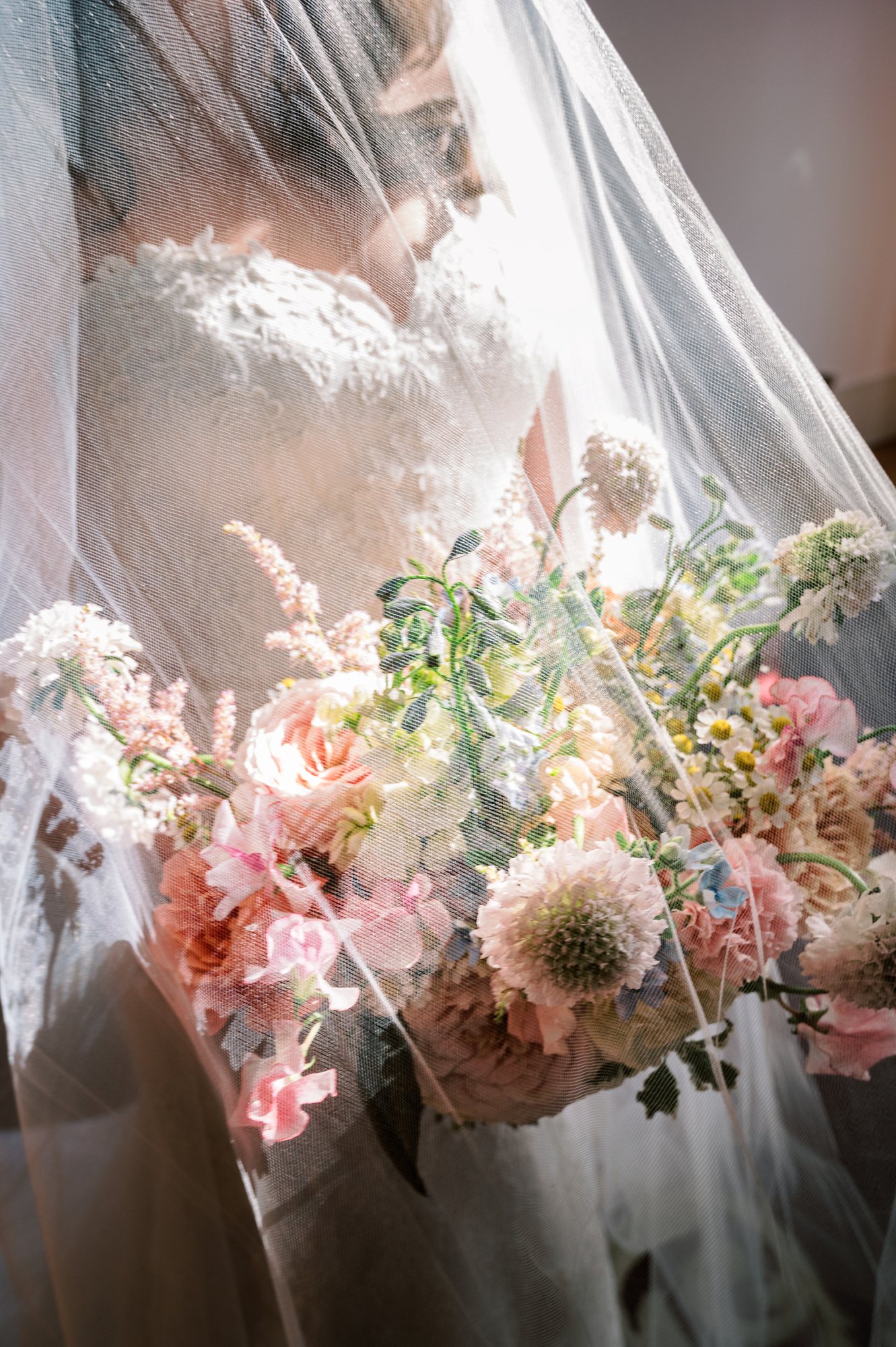 Bride beneath her veil holding a pastel bouquet in soft light